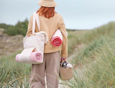 Woman in beige pants and a tan jacket with matching hat is carrying with a tote and yoga mat while walking through grass.
