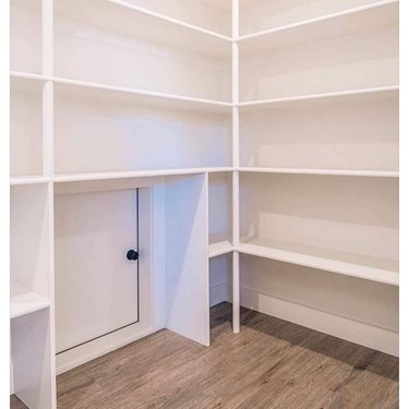 A white pantry with shelving above a small square door with a black doorknob.