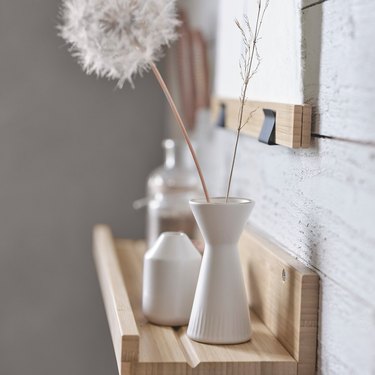 White vase with a puffy white flower next to a smaller white vase on a wooden shelf