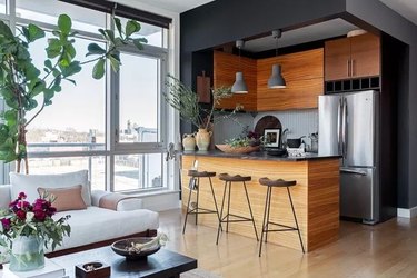 Modern kitchen with wood cabinets, black walls, wood floors, kitchen island, stools, plant.