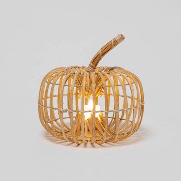 A pumpin lantern make of wood with a lit fake candle in the interior center of the pumpkin. It is on a light grey background.