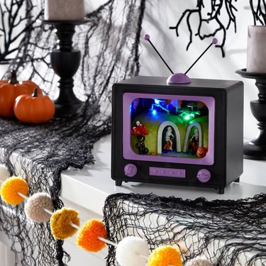 A small prop black and purple TV on a mantel with black spiderwebs, orange small pumpkins, and a pom-pom garland with white, orange, and beige coloring. There are two black candlesticks with white candles on either side of the TV. In the TV, you can see skeletons doing different activities, like riding a bike, listening to music, doing yoga, and hiding behind a bush.
