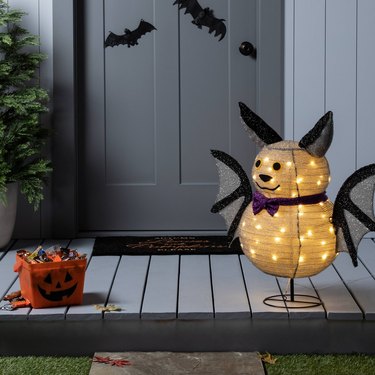An adorable light-up bat sculpture with a white paper lantern body and head, along with black wings, black ears, and a purple bowtie. The bat is on a light blue porch where there is also a pumpkin box filled with candy. On the front door, there are black bat decals.