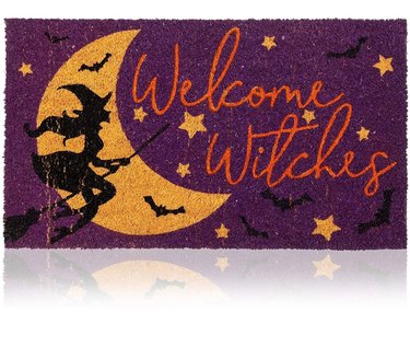 A purple doormat that has a yellow moon and stars, black bats, and a witch colored in black flying across the moon. It says "Welcome Witches" in an orange cursive font.