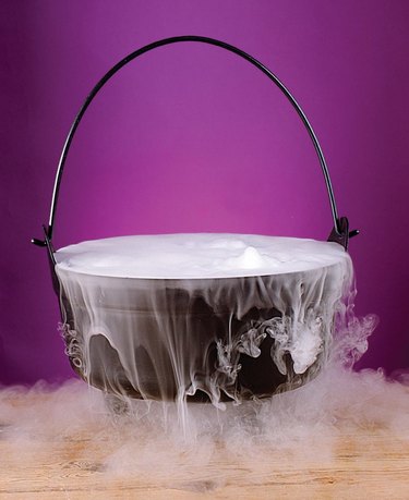 A black cauldron on a light wood table in front of a medium-purple background. The cauldron has white smoke spilling out of it from dry ice.