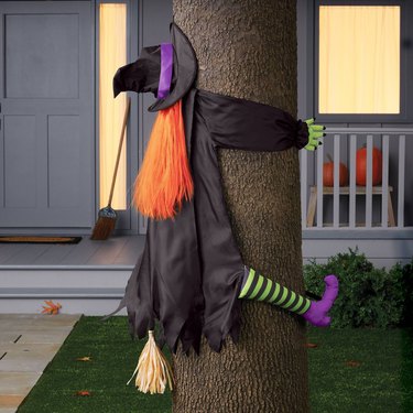 A fake witch crashing into a tree. The witch has a black cloak, black and green striped socks, purple heeled shoes, orange hair, a straw broom, and a black witch hat with a purple ribbon. Behind the tree, there is a light blue-gray porch with pumpkins and a broom on it.