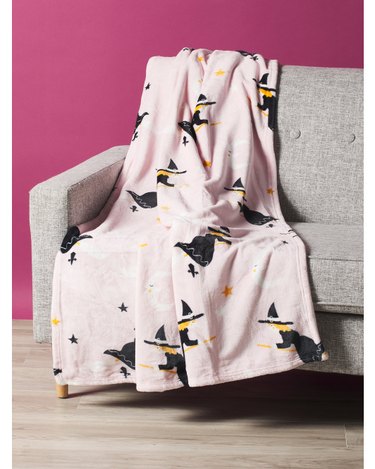 A pink fluffy throw blanket on a light grey fabric couch in front of a magenta wall. The pink blanket has flying witches, stars, and moons all over it.
