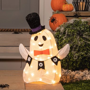 On a walkway, there is a white ghost sculpture with a string sign that says "boo." The ghost is wearing an orange bowtie and black top-hat with a purple ribbon. The porch is behind the ghost with orange pumpkins and black fake spiders on it. There is also a green bush covered in spiderwebs in the shot.