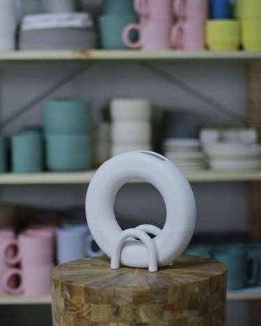 Round, white vase on a wooden platform in front of a shelf full of mugs/bowls