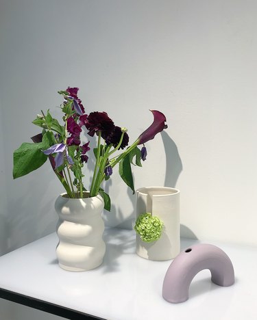 A white flower vase with flowers, a while cup, and a purple vase shaped like a rainbow on a white table