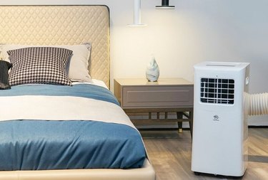 White portable air conditioner in beige and white bedroom with blue and white bed linens and black accents