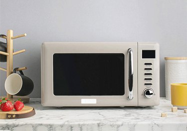 A beige microwave on a marble countertop with a mug holder and a plate of strawberries.