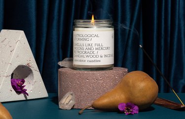 The Astrological Storming candle in a clear jar on a circular rock next to a yellow pear, clear crystal, purple violets, and burning incense stick.
