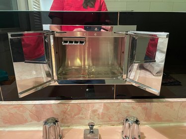 A tiny mirrored storage cupboard beneath a bathroom mirror and above a pink piled sink.