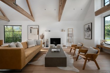 A Scandinavian living room with a light brown coach, two wood chairs, a gray coffee table, and a white fireplace that blends with the white walls.