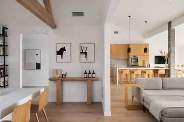 A living room, dining room, and kitchen decorate in a neutral Scandinavian palette with medium wood cabinets and chairs.