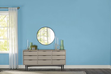 A beige dresser with a round mirror in front of a blue wall with a window off to the side.