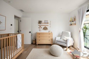 A Scandinavian nursery with a wood crib and dresser, a white armchair, a knit pouf on the floor, and various pieces of art and books shelved on the white walls.