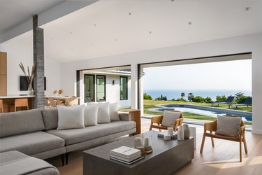 A Malibu living room with a couch and two wood chairs, plus two large open windows that face the ocean and an in-ground pool.