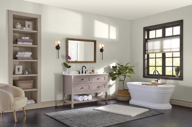 A bathroom with beige walls, a tub, vanity, mirror, beige shelving, and a beige chair in the corner