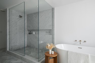 A white bathroom with a white and gray marble standing shower surrounded by glass, a white soaking tub, and a wood side table topped with a white vase of beige dried flowers.