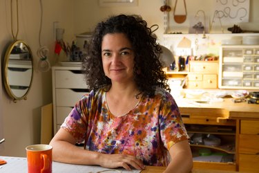 Sol Proaño, a white Latina woman, sits in her art studio. She has dark brown curly hair, dark brown eyes, and is wearing a white, pink, orange and purple short sleeved shirt. She sits at a table next to an orange coffee mug, and her tools are organized at a desk behind her.