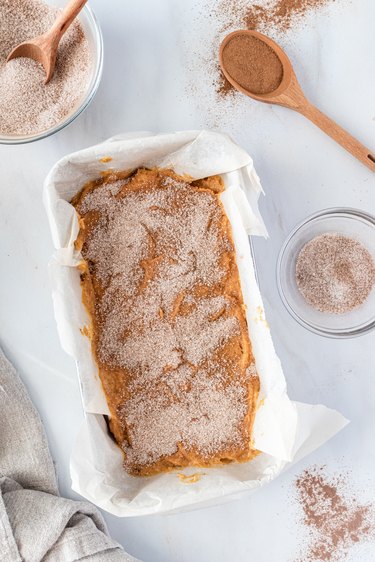 A loaf pan lined with parchment paper and filled with the batter that is topped with cinnamon-sugar.