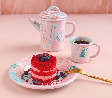A blue and pink swirled pitcher, mug, and plate with red cake with blueberries on top and a gold fork.