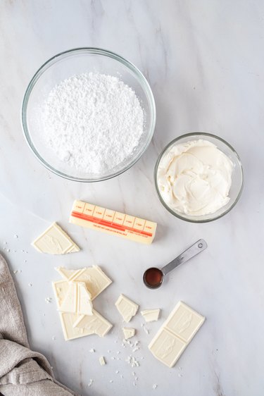 Glass bowls filled with powdered sugar and cream cheese, next to a stick of butter, teaspoon of vanilla extract, and pieces of white chocolate on a white marble countertop.