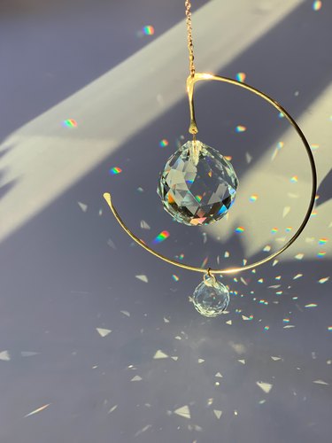An image of Sol Proano’s suncatcher, Prisma 3. It is a curved brass wire hanging, in the shape of an incomplete circle, with two clear crystal prisms hanging from it. The prisma hangs in front of a white wall, splashed with sun beams and small flecks of light reflecting off of the crystals.