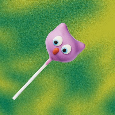 Starbucks Owl Cake Pop, a bite-sized piece of vanilla cake with buttercream dipped in a purple chocolate icing with an owl design in front of a green and yellow background.