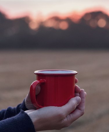 A person holding a red enamel mug outside while watching the sunset in a field.
