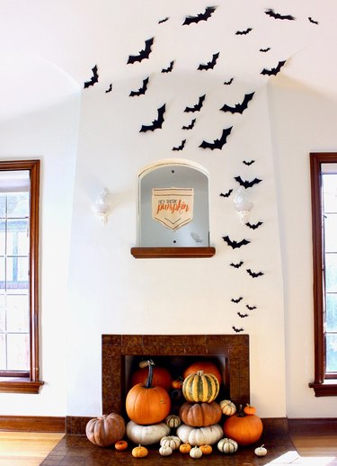 A white fireplace with paper bats that look like they are flying out of it. The hearth is filled with pumpkins of different shapes, sizes, and colors.
