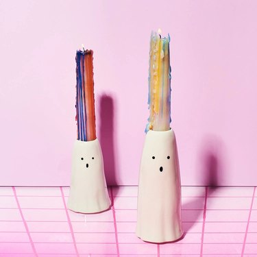 Two tall tapered candle holders in the shape of a ghost. Each has a melted multicolored candle in it. They are on a background with a pink wall and pink checked floor.
