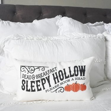 A bed covered in white pillows. The front throw pillow says "Dead and Breakfast. Sleepy Hollow. Please book a head." Below, there are three orange pumpkins. Around the text, there is black filigree.