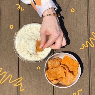 Hand dipping an orange chip into a creamy, white dip on a wooden table