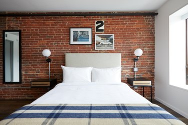 Industrial bedroom with brick wall, upholstered headboard, globe sconces, nightstands, mirror.