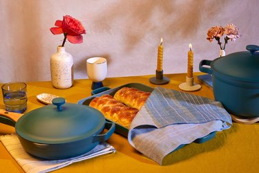 The Our Place's Shabbat Set including a teal oven pan with challah covered by a cloth, two candlesticks, an oven mat, a vase with a pink flower all on a wooden table
