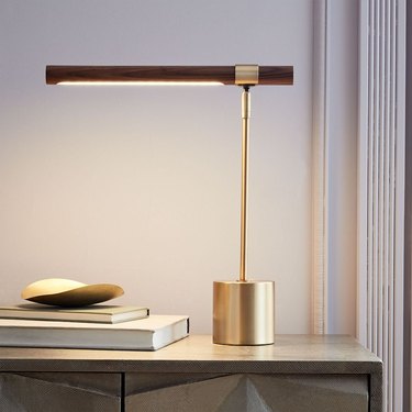 A brass linear piano lamp shining light on a table next to a small stack of books.