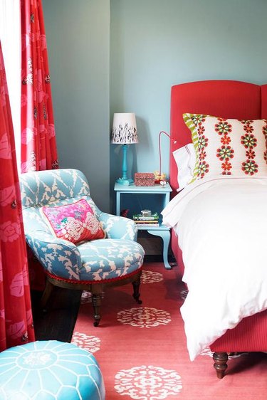 Bedroom with bold red bed frame, curtains, and rug, light blue chair, nightstand, and ottoman, and gray walls.
