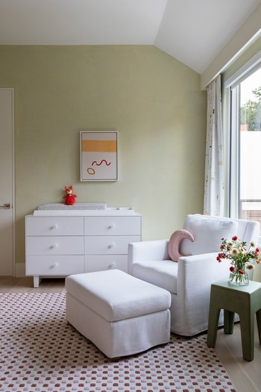 A light green nursery with white chair, white dresser, and red carpet and accents
