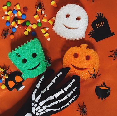 Three sponges shaped like an adorable Frankenstein, ghost, and pumpkin sponge. There are surrounded by candy corn and multicolored M&Ms. A skeleton hand is reaching toward the pumpkin sponge.