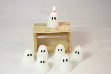 Seven tiny candles shaped like ghosts with ovular eyes. There is a small wood pallet behind six of the ghosts, and one has been lit and placed on top of the pallet. The background is white.