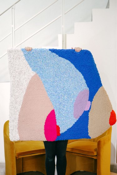 Colorful rug of three breasts and nipples being held by two hands with white staircase and rust chairs in the background