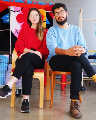 A woman with long brown hair and long-sleeved red sweater and a bearded man with short brown hair and light blue sweater both wear glasses and black jeans sit next to each other on different chairs, with opposite legs crossed over their knees.