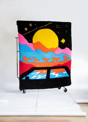 A rectangular carpet depicting a road trip scene with a black car and multicolored mountain scape with orange horses and a large moon with stars in a pitch-black sky.