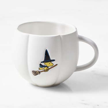A white pumpkin-shaped mug with the Peanuts character Woodstock (a yellow bird) flying on a broomstick while wearing a witch's hat and cape.