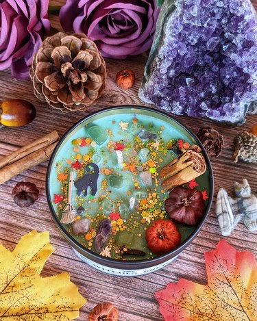 A Tiffany-blue candle with glitter, mini pumpkins, crystals, a black cat cutout, and a broom on top of it. It is surrounded by an amethyst, maple leaves, a pinecone, and a cinnamon stick.