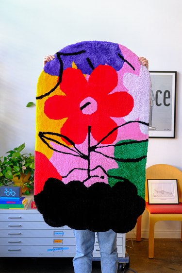 Person is covered holding multicolored patterned oval-like rug with red daisy in the middle and black leaves and dirt 