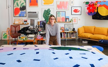 Woman with long brownish blonde hair and black rimmed glasses stands in front of a large confetti rug with a white wall of different framed artwork .